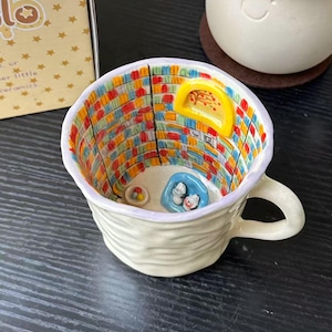 Puede incluir: Una taza de cer&aacute;mica con exterior color crema texturizado y un interior pintado a mano que muestra una escena de biblioteca colorida con estanter&iacute;as, una ventana arqueada amarilla, un sill&oacute;n azul peque&ntilde;o con dos figuras blancas y un cuenco.