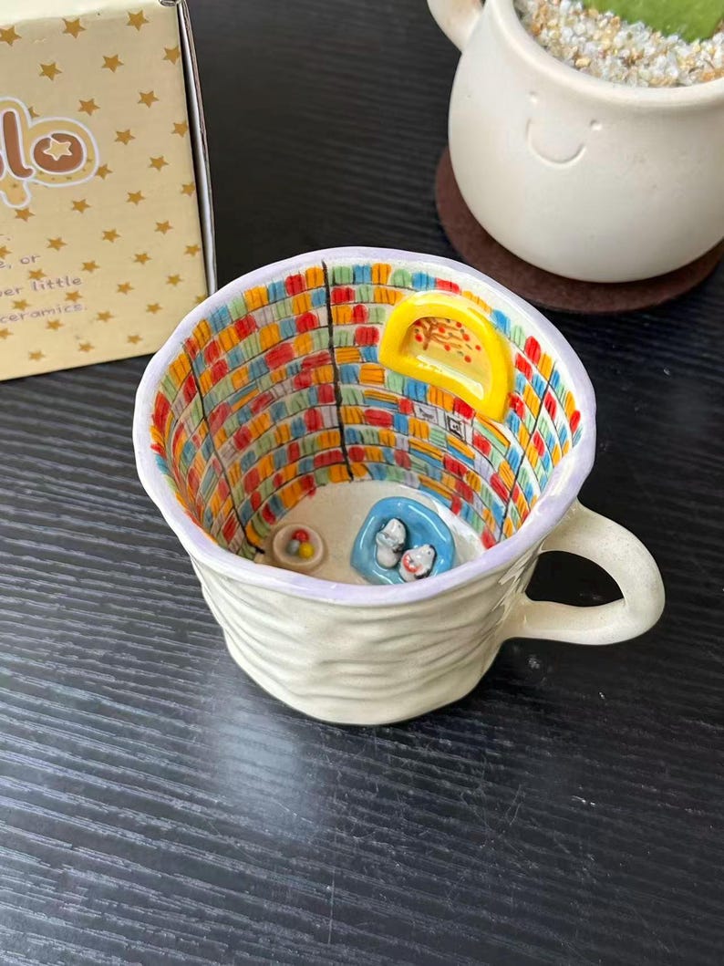 Puede incluir: Una taza de cer&aacute;mica con exterior color crema texturizado y un interior pintado a mano que muestra una escena de biblioteca colorida con estanter&iacute;as, una ventana arqueada amarilla, un sill&oacute;n azul peque&ntilde;o con dos figuras blancas y un cuenco.