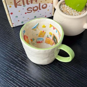 Op de afbeelding: Een crèmekleurige keramische mok met een lichtgroene rand en handvat. De binnenkant is voorzien van verhoogde, kleurrijke miniatuursculpturen van schoolspullen, waaronder boeken, een schoolbord en een papieren vliegtuigje. Achter de mok staat een doos.