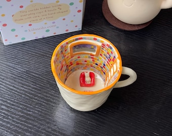 Può includere: Una tazza in ceramica con esterno bianco testurizzato e bordo arancione. L'interno presenta una scena di biblioteca dipinta a mano con scaffali colorati, una finestra e una piccola sedia rossa tridimensionale con una statuetta di gatto bianco.