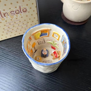 以下が含まれることがあります： 青い縁取りが施された陶器製のカップで、内部には3Dのミニチュアの部屋の風景が描かれています。内壁には小さな額縁に入ったアートが飾られ、底面には椅子に座った小さなフィギュア、テーブル、鉢植えが配置されています。