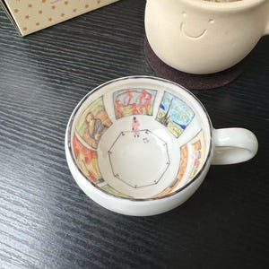 Könnte beinhalten: Eine weiße Teetasse aus Keramik mit einer handgemalten Illustration einer Kunstgalerie im Inneren. Das Kunstwerk zeigt verschiedene gerahmte Gemälde an den Wänden und eine kleine Figur, die in der Mitte des Raumes steht. Die Tasse hat einen Henkel.