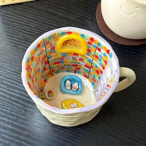 Puede incluir: Una taza de cer&aacute;mica que presenta una escena interior en miniatura de una biblioteca con estanter&iacute;as coloridas, una ventana arqueada amarilla, un sill&oacute;n azul con dos figuras peque&ntilde;as, una alfombra amarilla con un libro y un cuenco peque&ntilde;o.