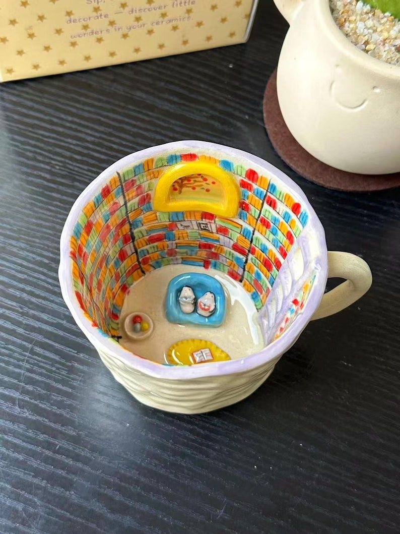 Puede incluir: Una taza de cer&aacute;mica que presenta una escena interior en miniatura de una biblioteca con estanter&iacute;as coloridas, una ventana arqueada amarilla, un sill&oacute;n azul con dos figuras peque&ntilde;as, una alfombra amarilla con un libro y un cuenco peque&ntilde;o.