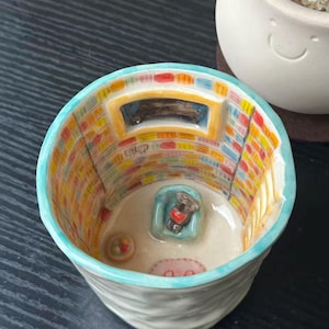 May include: A ceramic cup featuring an interior diorama of a room with a small koala figurine sitting in a light blue chair, a rug, and a bowl. The walls are painted with colorful rectangular patterns and a window, with a light blue rim.