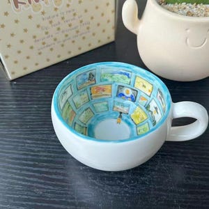 Puede incluir: Una taza de té de cerámica blanca con asa, que presenta un diseño pintado a mano en el interior de una pared de galería con múltiples pinturas de paisajes enmarcadas en tonos azules, amarillos y verdes, y una pequeña figura en el centro.