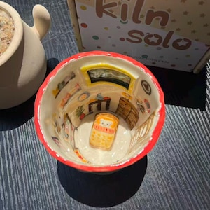 Peut inclure: Un mug en céramique avec un bord rouge vif et une scène intérieure peinte à la main représentant une pièce avec un petit lit, des meubles et des décorations murales. Le mug est placé devant une boîte en carton portant le texte kiln solo et small joys.