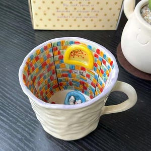 Puede incluir: Una taza de cer&aacute;mica con exterior texturizado en color blanco roto y un asa. El interior presenta una escena de biblioteca pintada a mano con estanter&iacute;as coloridas, una ventana amarilla arqueada y una silla azul con dos figuras blancas.