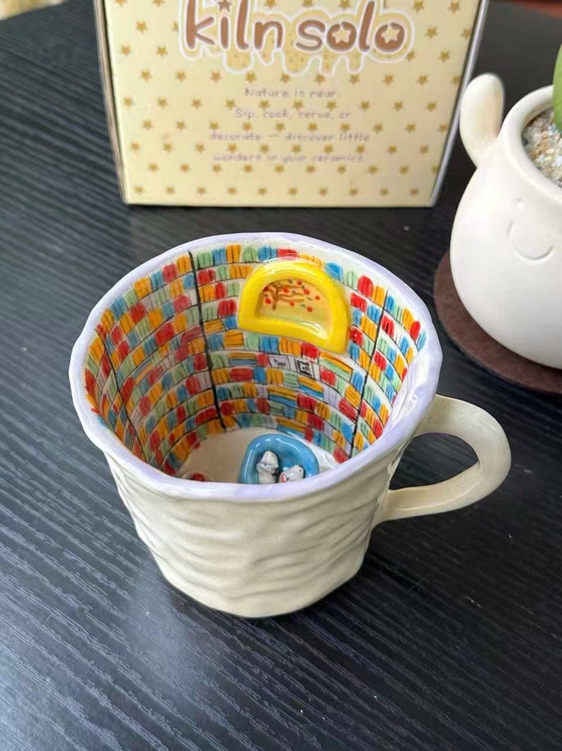Puede incluir: Una taza de cer&aacute;mica con exterior texturizado en color blanco roto y un asa. El interior presenta una escena de biblioteca pintada a mano con estanter&iacute;as coloridas, una ventana amarilla arqueada y una silla azul con dos figuras blancas.