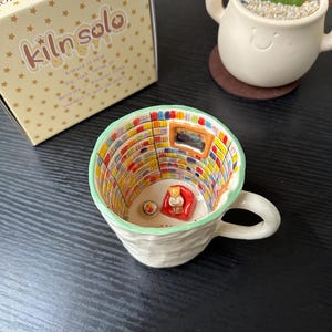 May include: A ceramic mug with a textured off-white exterior and a hand-painted interior featuring a miniature library scene with colorful bookshelves, a small orange armchair, and a tiny figure sitting inside, next to a box labeled Kiln Solo.