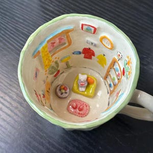 Può includere: Una tazza in ceramica con interno dipinto a mano che presenta una scena in miniatura in 3D di una stanza. Le pareti interne mostrano finestre, vestiti e decorazioni dipinte, mentre la base contiene un piccolo divano giallo con una figura bianca, un tappeto e un tavolino.