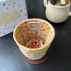 May include: A ceramic cup with a textured off-white exterior and a detailed interior featuring a miniature library scene. The interior walls show colorful bookshelves, a ladder, and a small orange pet bed with two tiny figures on a floor.