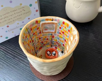 Puede incluir: Una taza de cerámica con un exterior texturizado en color blanco roto y un interior detallado que muestra una escena de biblioteca en miniatura. Las paredes interiores presentan estanterías coloridas, una escalera y una cama naranja.