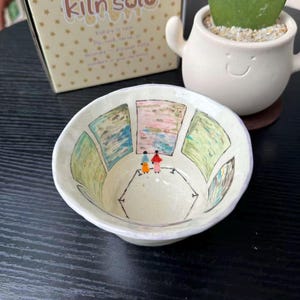 May include: A small, off-white ceramic bowl featuring a hand-painted interior scene of two figures standing in an art gallery viewing framed landscape paintings. The bowl is positioned in front of a cardboard box labeled kiln solo and a small white planter.