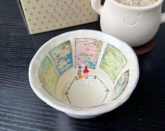 Può includere: Una piccola ciotola in ceramica color bianco sporco con una scena dipinta a mano all'interno che raffigura due figure in una galleria d'arte mentre osservano dei dipinti di paesaggi incorniciati. La ciotola è posta davanti a una scatola di cartone con la scritta kiln solo e un piccolo vaso bianco.