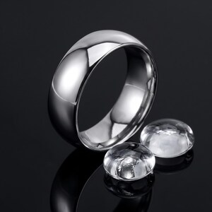 May include: A polished silver-toned metal ring with a smooth, rounded band and a high-gloss reflective finish. The ring is positioned next to two small, clear, rounded glass cabochons that also feature a smooth, reflective surface.
