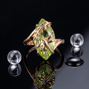 May include: A rose gold ring featuring a marquise-cut light green gemstone. The setting includes a metal dragonfly motif resting on the stone and a branch-like design extending along the side of the band. The ring is displayed with clear spheres.