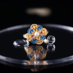 May include: A gold ring featuring a floral-shaped centerpiece with blue enamel detailing, a central white pearl, and small red gemstone accents. The intricate metalwork creates a filigree pattern, and the ring is displayed with clear glass spheres.