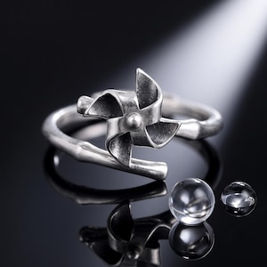 May include: A silver-toned ring featuring a pinwheel-shaped centerpiece and a band designed to resemble a bamboo stalk. The ring has an open-ended construction and is accompanied by two small, clear, spherical glass beads placed nearby.