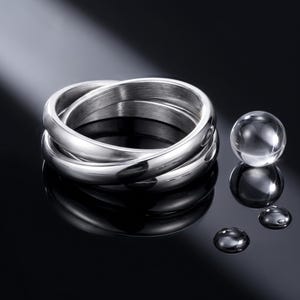 May include: A silver-toned triple-band interlocking ring with a polished metallic finish. Beside the ring are three clear, spherical glass beads of varying sizes, reflecting light on a dark, reflective surface with a smooth, glossy appearance.