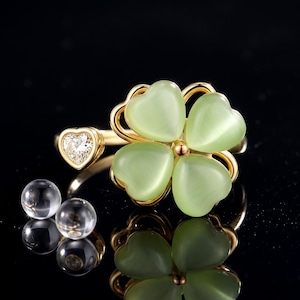 May include: A gold ring featuring a four-leaf clover design composed of four pale green heart-shaped cat's eye stones. One end of the open band is accented with a single small heart-shaped clear gemstone. Two small clear spheres sit nearby.