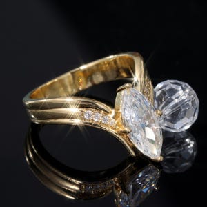 Könnte beinhalten: Ein goldfarbener Ring mit einem zentralen, marquise-geschliffenen, klaren Stein, der durch eine Reihe kleiner, runder, klarer Steine entlang der Ringschiene akzentuiert wird. Eine kugelförmige, facettierte Perle ist seitlich an der Fassung befestigt.