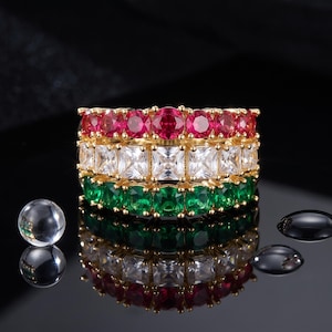 May include: A gold-toned ring featuring three horizontal rows of faceted stones. The top row consists of round red stones, the middle row features square white stones, and the bottom row contains round green stones, all set in a metallic band.