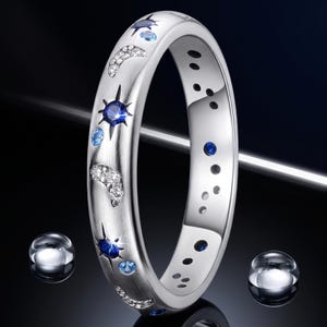 May include: A silver-toned ring featuring a brushed metal finish, decorated with inlaid celestial motifs including crescent moons paved with clear stones, dark blue star-shaped gems, and smaller light blue circular stones set along the band.