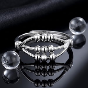 May include: A silver-toned ring featuring a design inspired by an abacus, with three horizontal bars each holding three small, movable silver beads. The ring has a polished, reflective finish and is accompanied by two clear, spherical glass beads.