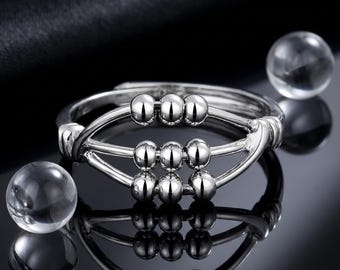 Puede incluir: Un anillo de tono plateado con un diseño inspirado en un ábaco, que presenta tres barras horizontales con tres pequeñas cuentas plateadas móviles cada una. El anillo tiene un acabado pulido y reflectante, junto a dos cuentas de vidrio esféricas.