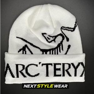 May include: A white knit beanie featuring a black graphic design of a stylized creature on the crown and the text ARC'TERYX printed in bold black capital letters across the folded cuff. The hat is constructed from a soft, textured knit fabric.