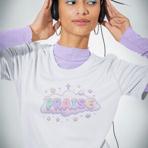Op de afbeelding: Een wit T-shirt met korte mouwen en een afbeelding van een lichtpaarse wolk met het woord PRAISE in pastelkleurige bubbelletters. Het ontwerp bevat kleine sterretjes, een kruisje en een hartje rondom de wolk, gedragen over een lavendelkleurig shirt met lange mouwen.