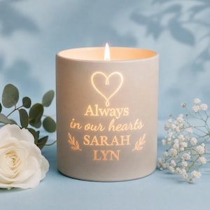 May include: A cylindrical light grey candle holder with a glowing interior. The front features a heart outline above the text Always in our hearts SARAH LYN, flanked by small leaf sprigs. The candle emits a warm light through the etched design.