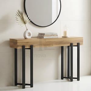 May include: A rectangular console table with a light brown wood grain top and a black metal frame consisting of four vertical legs on each side. The table surface holds a white ceramic vase with a dried palm leaf, three stacked books, and a candle.