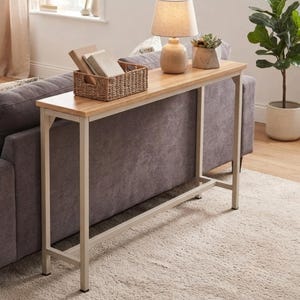 May include: A narrow console table featuring a light wood grain rectangular top and a sturdy off-white metal frame. The table holds a woven rectangular basket containing books, a beige table lamp with a fabric shade, and a small succulent plant.