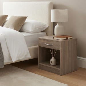 May include: A wooden bedside table with a light brown wood grain finish, featuring one drawer with a silver handle and an open storage shelf below. A white table lamp, a closed brown notebook, and a small white ceramic vase with sticks sit on top.
