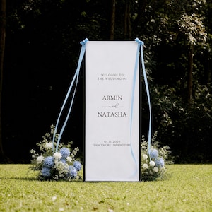 May include: A tall, rectangular white wedding welcome sign with a black frame, featuring the text "WELCOME TO THE WEDDING OF ARMIN and NATASHA 01.11.2026 LANCEMORE LINDENDERRY". Light blue ribbons are tied to the top corners, with blue and white floral arrangements at the base.
