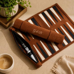 May include: A brown suede backgammon board featuring black and white triangular points. A matching brown suede roll-up case with the monogram G&B is placed on top. A black and white doubling cube and dice are visible inside the open side pocket.