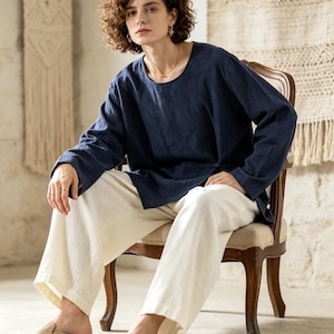 May include: A dark navy blue long-sleeved tunic with a round neckline and a small side toggle closure, paired with loose-fitting off-white trousers and beige suede slip-on shoes. The ensemble features a relaxed fit and a textured fabric appearance.
