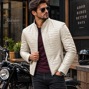 Puede incluir: Una chaqueta de color crema con un patrón texturizado de piel de cocodrilo, que presenta un cuello alzado, una cremallera frontal completa y mangas largas. La chaqueta se lleva sobre una camiseta de color burdeos oscuro y vaqueros gris oscuro.