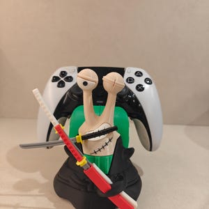 Può includere: Una figura beige simile a una lumaca con due occhi su steli, che indossa un indumento nero con una fascia verde, che tiene tre spade con else rosse, bianche e nere. La figura è posizionata davanti a un controller di gioco wireless bianco e nero.