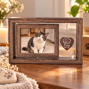 May include: A dark wood rotating picture frame containing a photograph of a brown and white tabby cat. A heart-shaped wooden pendant hangs from the frame, engraved with a cat illustration, the name Lucy, and the dates 2010-2024.