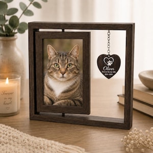 Puede incluir: Un marco de fotos giratorio de madera marrón oscuro que contiene una fotografía de un gato atigrado marrón. Un adorno de madera en forma de corazón cuelga del marco mediante una cadena metálica, grabado con una huella, el nombre Oliver y el texto Forever by my side 2011-2024.