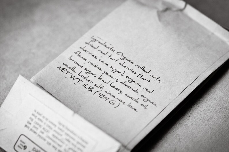May include: A close-up of a brown paper bag with a handwritten list of ingredients for a baked good. The ingredients include organic rolled oats, dried red tart cherries, organic red flame raisins, pecans, almonds, organic brown sugar, local honey, canola oil, vanilla, kosher salt, and cinnamon. The bag also includes the net weight of the product, 1 lb (454 g).