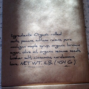 May include: A close-up of a brown paper bag with handwritten text listing ingredients for a food product. The ingredients include oats, pecans, maple syrup, and sesame seeds. The net weight is 1 lb (454 g).