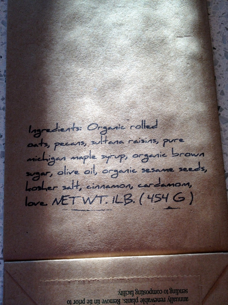 May include: A close-up of a brown paper bag with handwritten text listing ingredients for a food product. The ingredients include oats, pecans, maple syrup, and sesame seeds. The net weight is 1 lb (454 g).