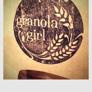 May include: A circular logo with the words "granola girl" in white, set against a dark, textured background. The logo includes wheat stalks and decorative flourishes, all on a tan background.