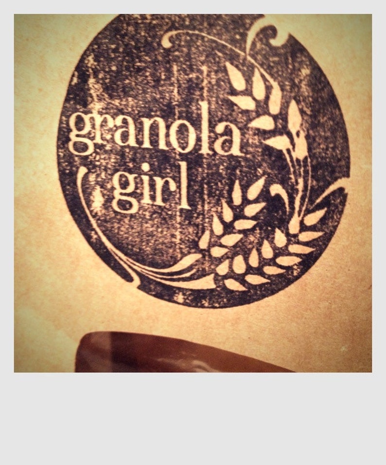 May include: A circular logo with the words "granola girl" in white, set against a dark, textured background. The logo includes wheat stalks and decorative flourishes, all on a tan background.