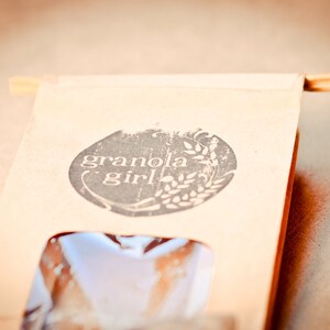 May include: A brown paper bag with a white logo that says "granola girl" in a circle with a wheat stalk design. The bag has a clear window showing the product inside.