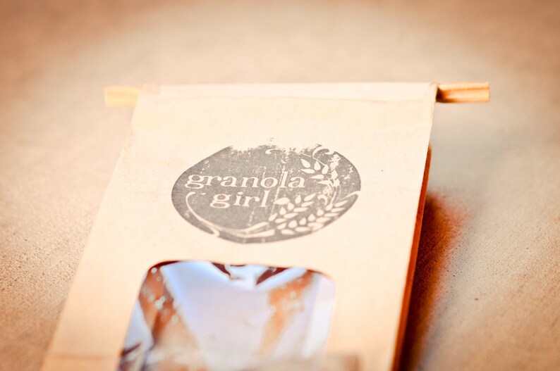 May include: A brown paper bag with a white logo that says "granola girl" in a circle with a wheat stalk design. The bag has a clear window showing the product inside.